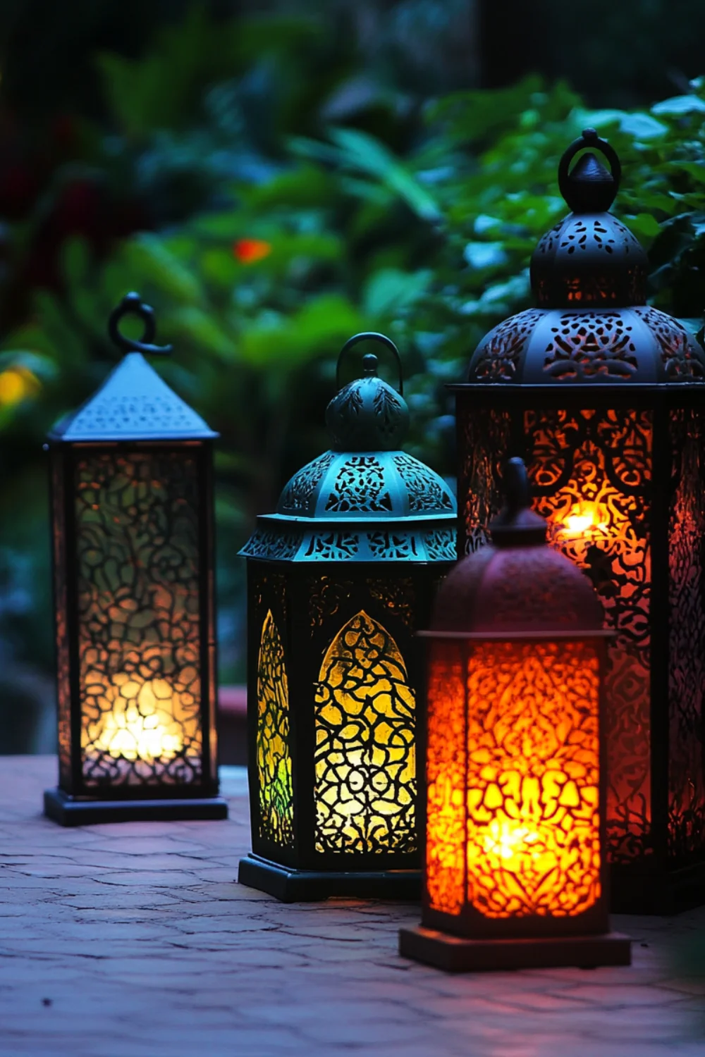 A set of ornate metal lanterns glowing with warm candlelight, placed on a rustic outdoor table with lush green foliage in the background.